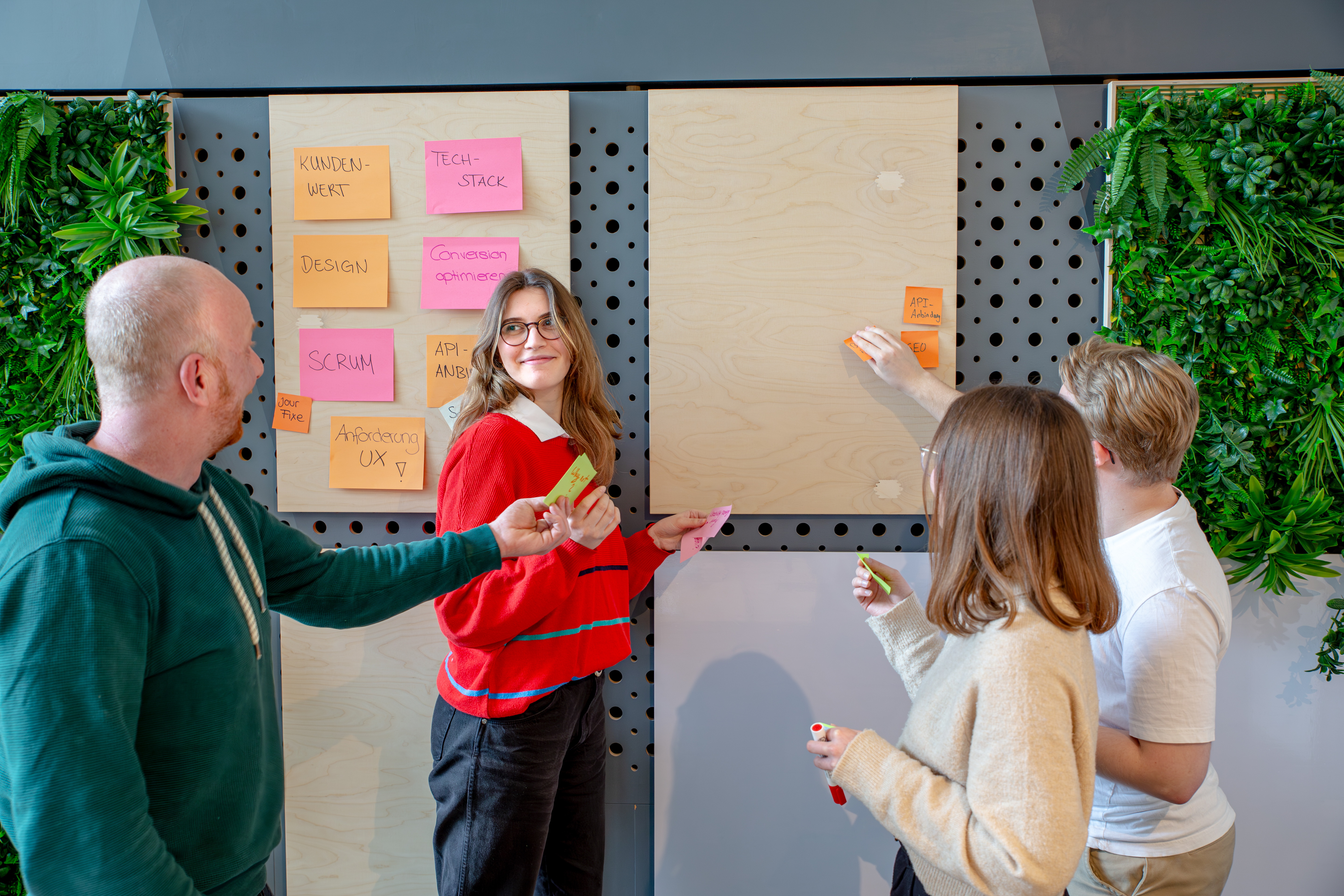 Vier Designer:innen stehen an einem Whiteboard und brainstormen gemeinsam. Die Person im Vordergrund reicht einem anderen Designer:in ein Post-it.